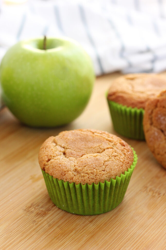 muffin in a green liner sitting on a wooden cutting board with two muffins and a green apple sitting off to the side