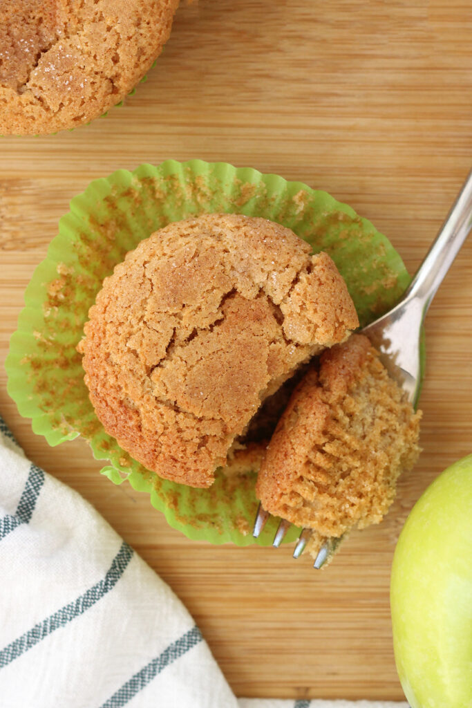 top down image of a muffin that is sitting on an open green liner with a bite sitting on a fork on a wooden tabletop