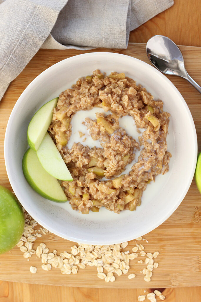 top down image showing a white bowl that is filled with oatmeal and green apples. The bowl is sitting on a wooden table top with additional uncooked oats spread around with green apples, a tan napkin, and a spoon off to the side