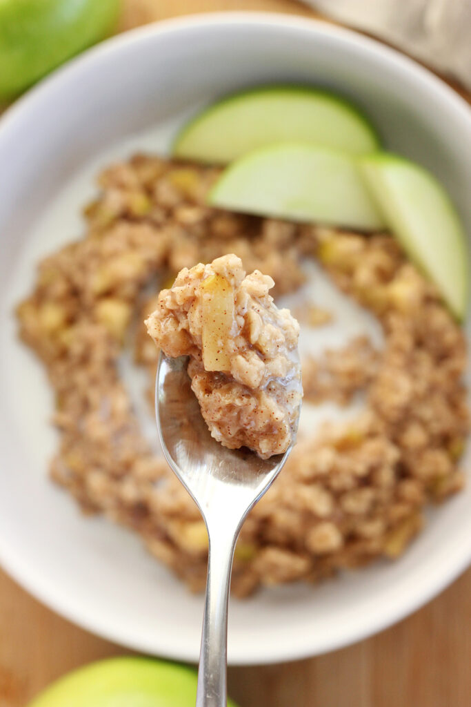 spoon holding a bite of oatmeal that has an apple piece on top. The spoon is being held over a shallow white bowl with more oatmeal and green apple slices. 