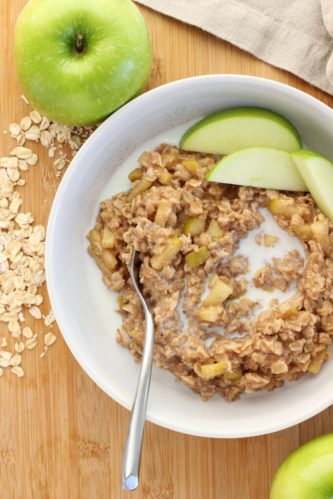 top down image of a  shallow white bowl with oatmeal and green apple slices. A spoon is in the oats with milk. 