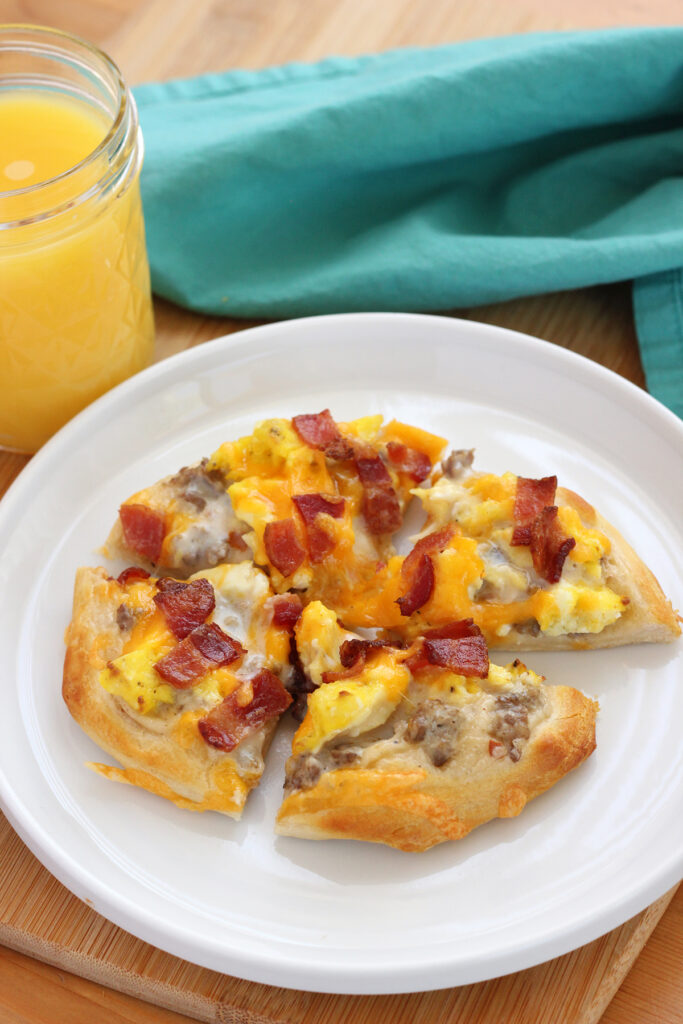 small white round plate with a lifted edge topped with a mini pizza that is topped with scrambled eggs, cheese and bacon sitting on a wooden cutting board with a glass of orange juice and teal napkin off to the side