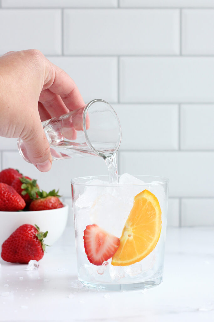 short glass filled with ice and a slice of orange and sliced strawberries with vodka being poured into it from a shot glass. A small white bowl of strawberries in the background