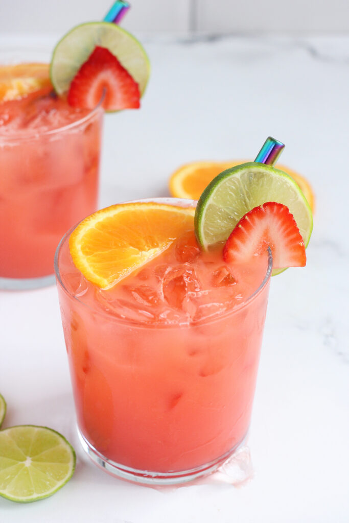close up on a short glass that is filled with a pink drink topped with an orange wedge, lime wedge, and strawberry slice with a glass of juice behind on a white background