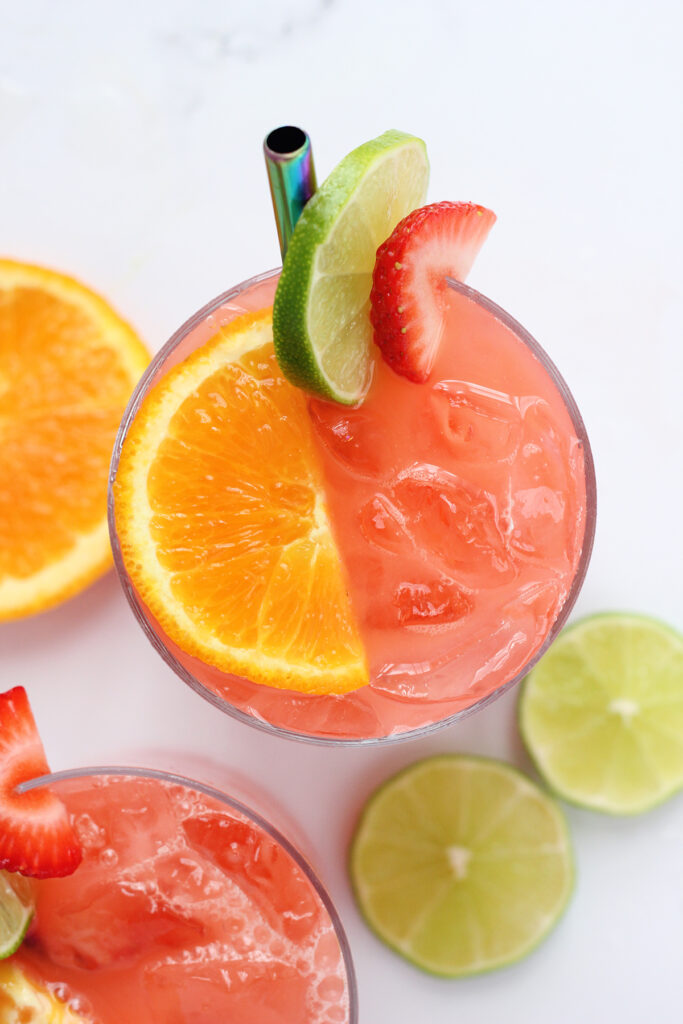 top down image of a glass filled with pink liquid and topped with an orange slice, strawberry slice, and lime edge. More fruit is laying off to the side.