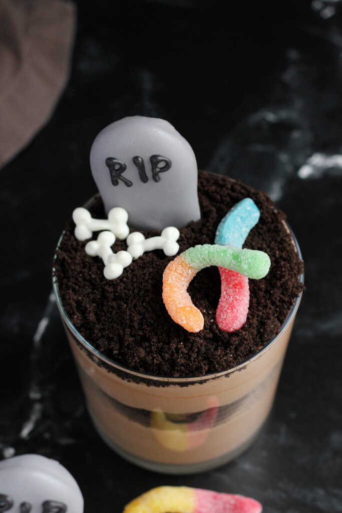 close up of a glass cup that is filled with layers of chocolate pudding and crushed cookies and decorated to look like a graveyard with cookies that appear to be gravestones and ghosts with little bone candies off to the side.
