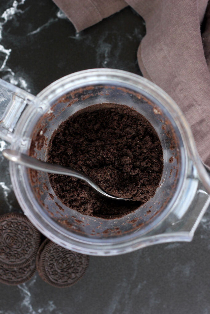 top down image showing the inside of a blender filled with cookie crumbs and a spoon. Additional cookies and a dark brown napkin sitting off to the side