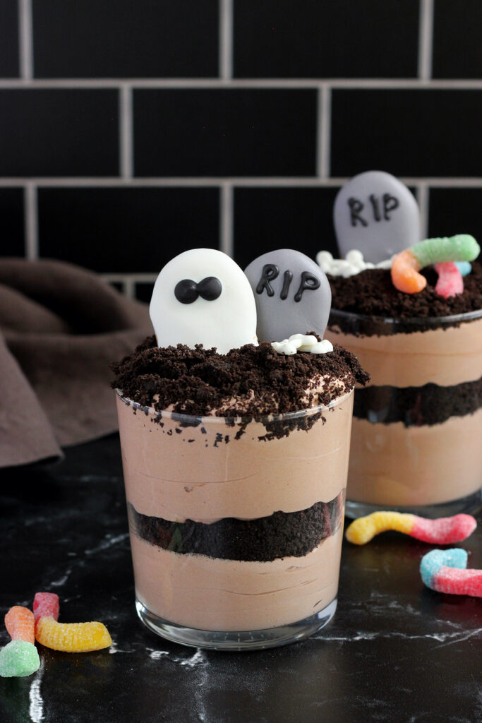close up of a glass cup that is filled with layers of chocolate pudding and crushed cookies and decorated to look like a graveyard with cookies that appear to be gravestones and ghosts with little bone candies off to the side.