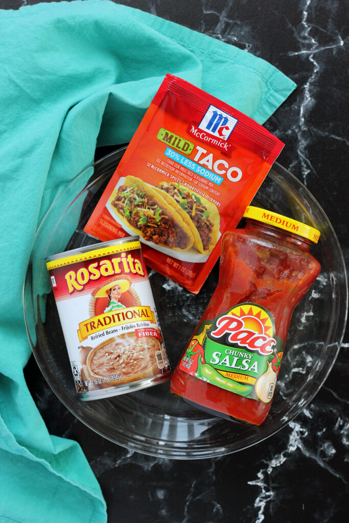 package of McCormick mild taco seasoning, can of Rosarita traditional beans, and a jar of PAce chunky salsa sitting on a shallow glass pan