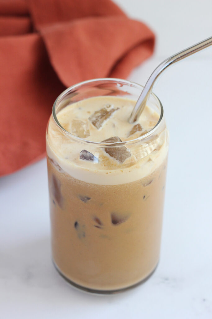 close up of a tall glass filled with an iced coffee with a metal straw sitting on a white counter with a burnt orange napkin off to the side