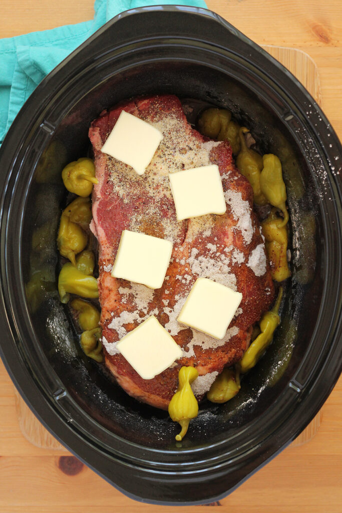 top down image of a slow cooker bowl filled with a roast, yellow peppers, seasonings, and dotted with butter sitting on a wooden table top with teal napkin