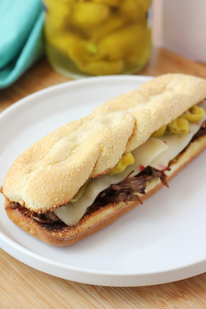 sub style sandwich roll filled with shredded beef, cheese, and sliced pepeprs sitting on a white plate that is on a wooden table top with a jar of peppers sitting in the background