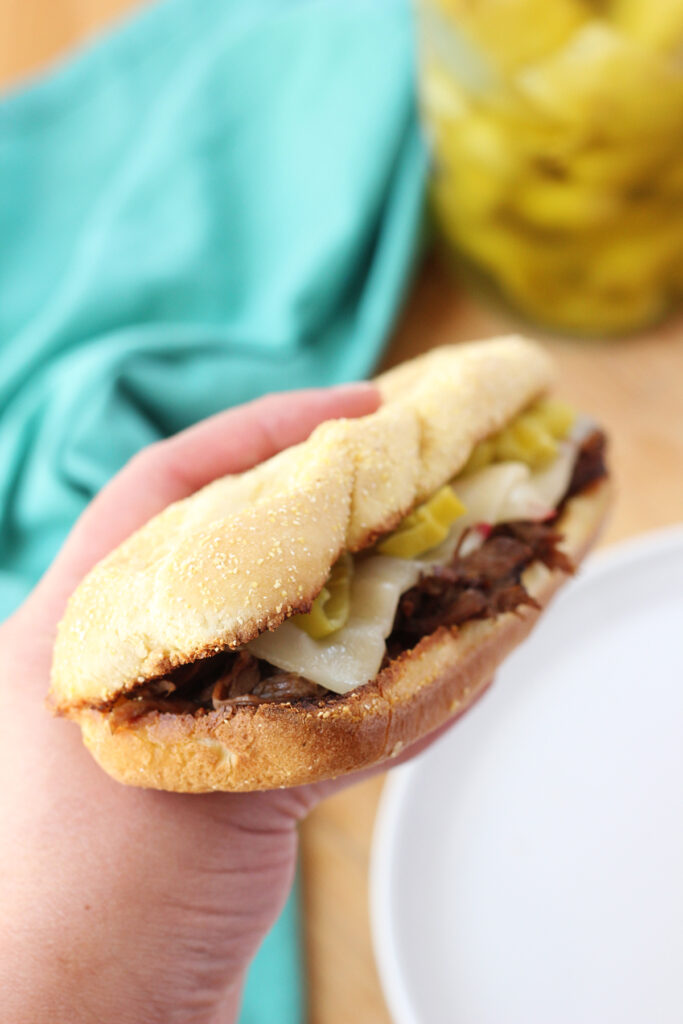 hand holding a sub style sandwich that is loaded with shredded beef, cheese and yellow peppers over a white plate and teal napkin