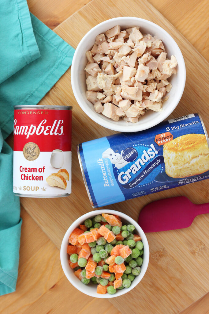 top down image showing a wooden cutting board of a white bowl filled with cooked chopped chicken, a can of Campbell's cream of chicken soup, a can of Grands southern homestyle biscuits,  and a smaller white bowl filled with frozen carrots and peas