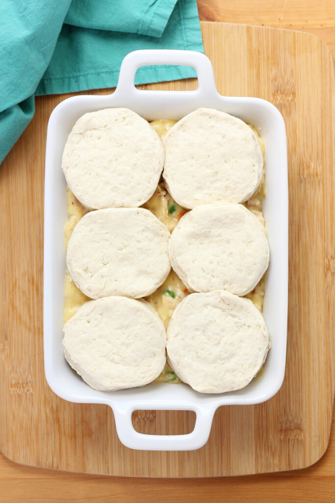 unbaked canned biscuits sitting on top of a chicken filling in a white rectangle baking dish. The dish is sitting on a wooden table top with a teal napkin off to the side