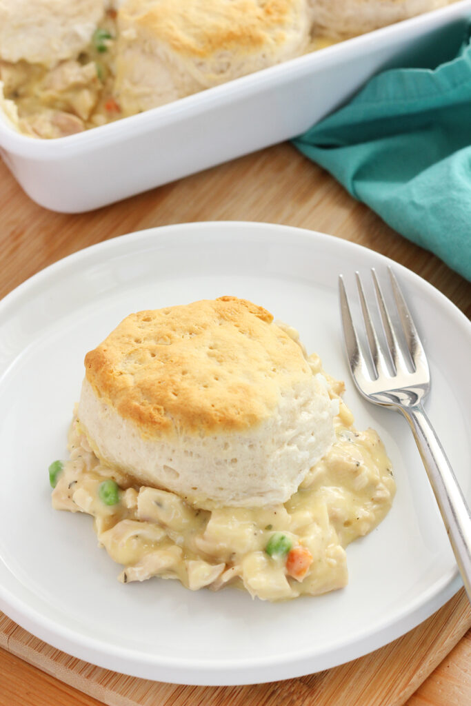 small white round plate with a slightly lifted edge topped with what appears to be pot pie filing topped with a biscuit. A fork is sittign off to the side with a baking dish with more of the bake is in the background with a teal napkin