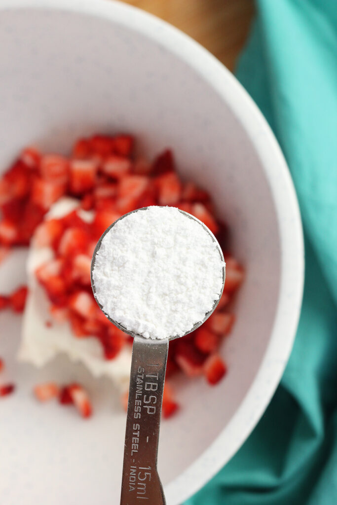 single tablespoon of powdered sugar being held over a bowl of chopped strawberries and a block of cream cheese with a teal napkin off to the side