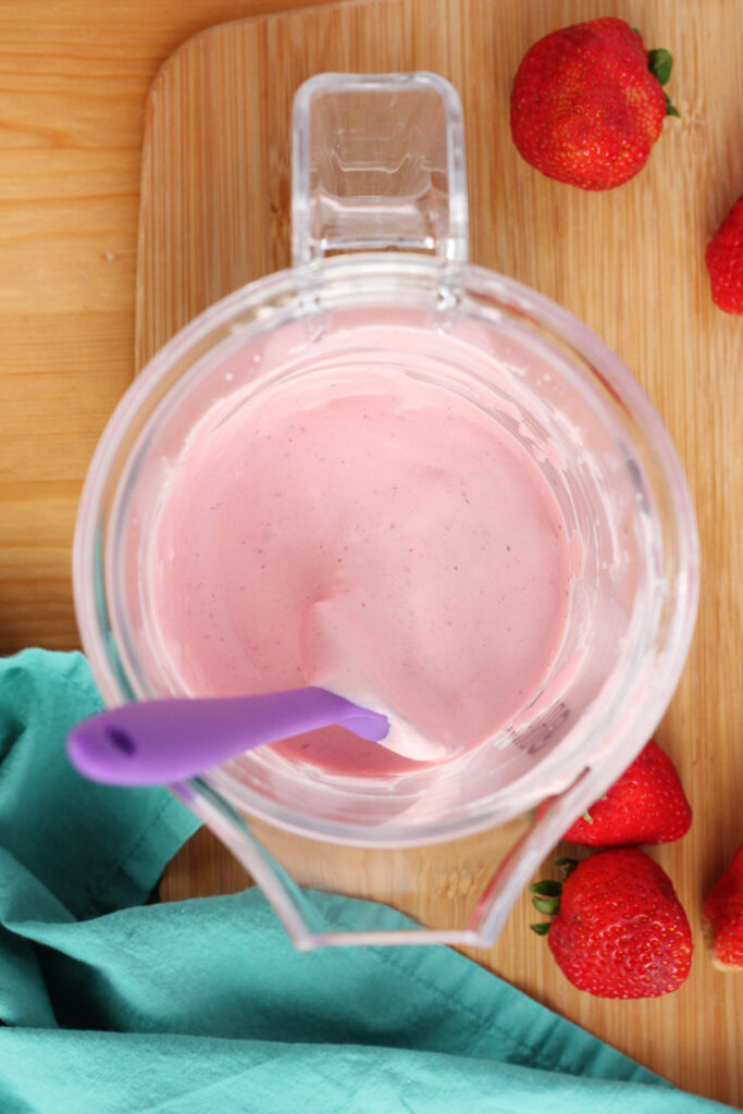 Top down shot of a blender filled with a pink liquid sitting on a wooden table top with a teal napkin and a purple spatula