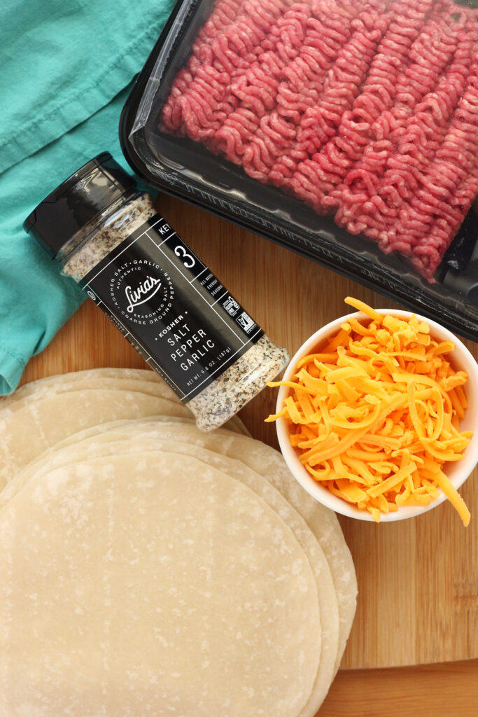 top down image showing a wooden cutting board that is topped with a teal napkin, uncooked ground beef, small white bowl of shredded cheese, stack of uncooked tortillas, and a container of Livias seasoning