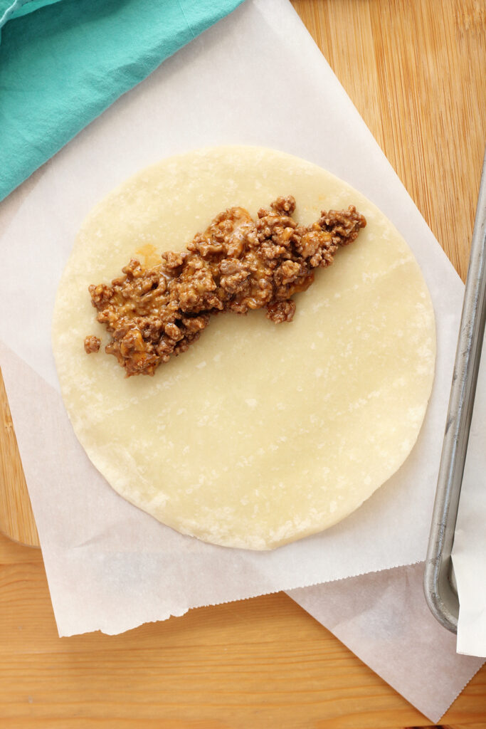 piece of parchment paper sitting on a wooden cutting board with a raw tortilla on top with a row of cooked ground beef to one side. 