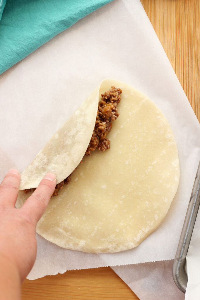 piece of parchment paper sitting on a wooden cutting board with a raw tortilla on top with a row of cooked ground beef to one side with a hand folding it over