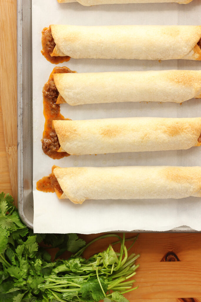 top down image showing a baking tray that is lined with taquitos. Tray is sitting on a wooden table top with cilantro off to the side