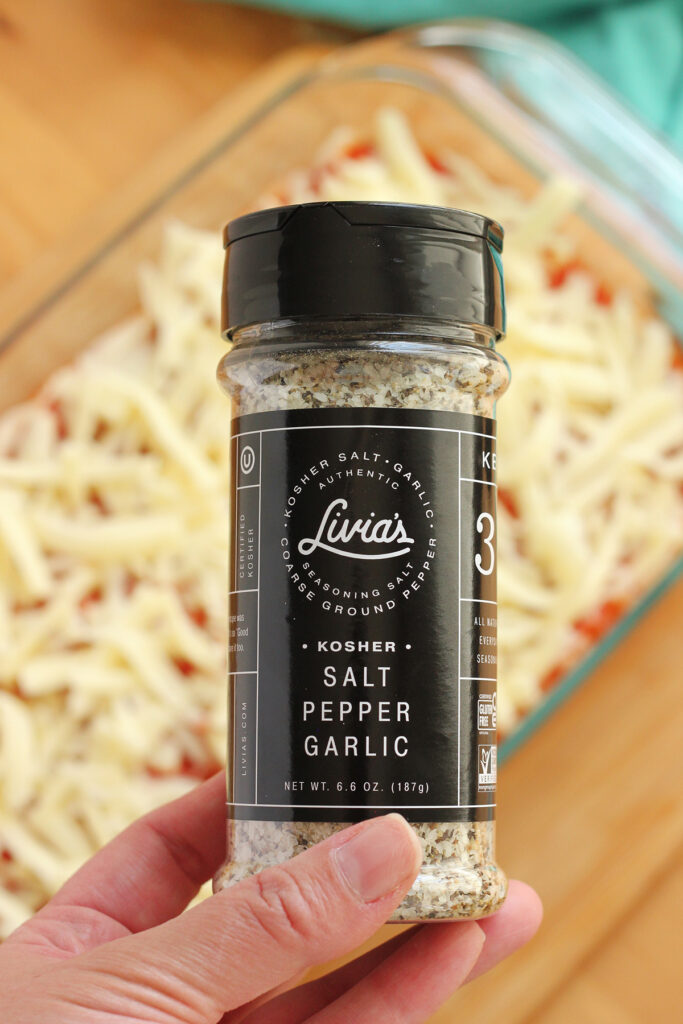 container of Livia's salt pepper and garlic mix being held over the top of an unbaked casserole