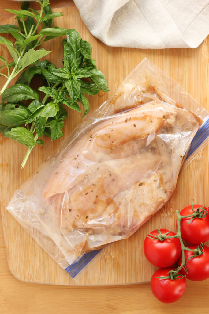 top down image showing a wooden table top that has fresh basil, tomatoes on a vine, and a bag filled with chicken breast in a marinade. A tan napkin is off to the side