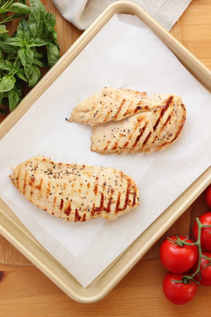 top down image showing a baking tray lined with parchment paper with two grilled chicken breast on top. Tray is sitting on a wooden table top with fresh basil and vine tomatoes off to the sides. 