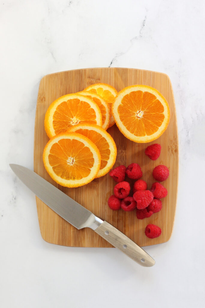 top down image showing a wooden cutting board with six orange slices and 13 raspberries sitting with a small knife on top of a white coutner.