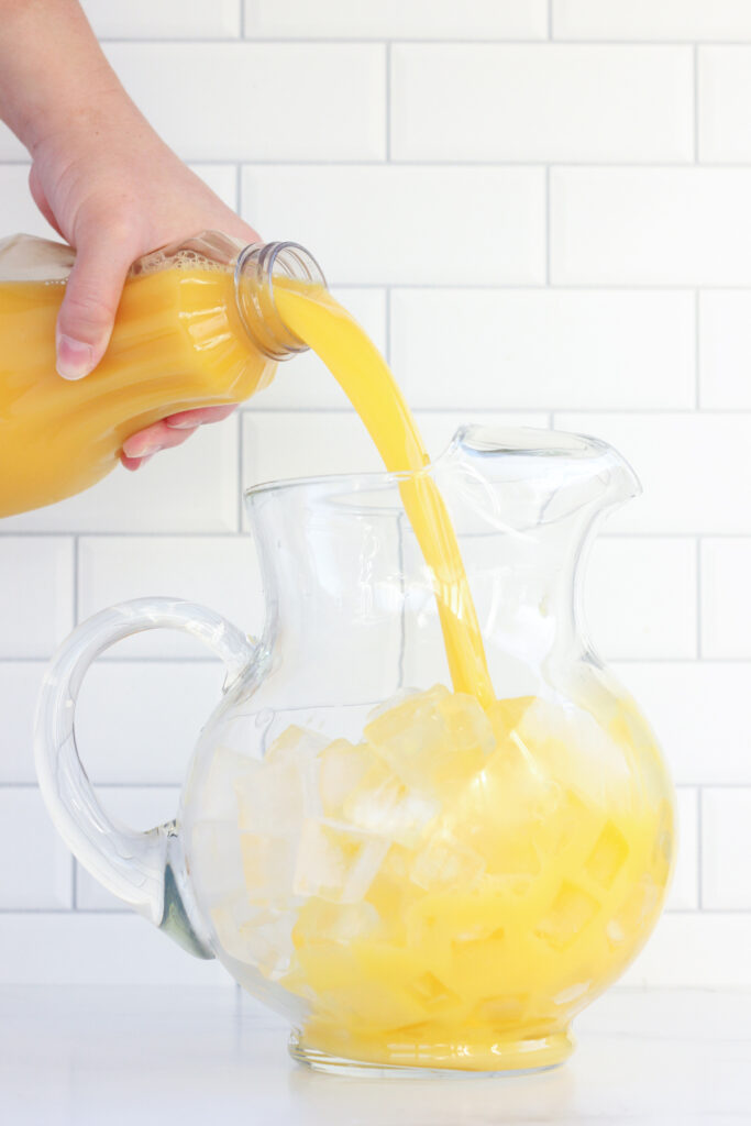 hand pouring orange juice from a container into a short pitcher filled with ice
