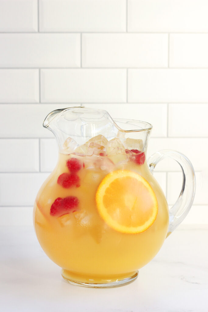 pitcher filled with ice and a drink mix that is yellow with a sliced orange and raspberries on a white table top