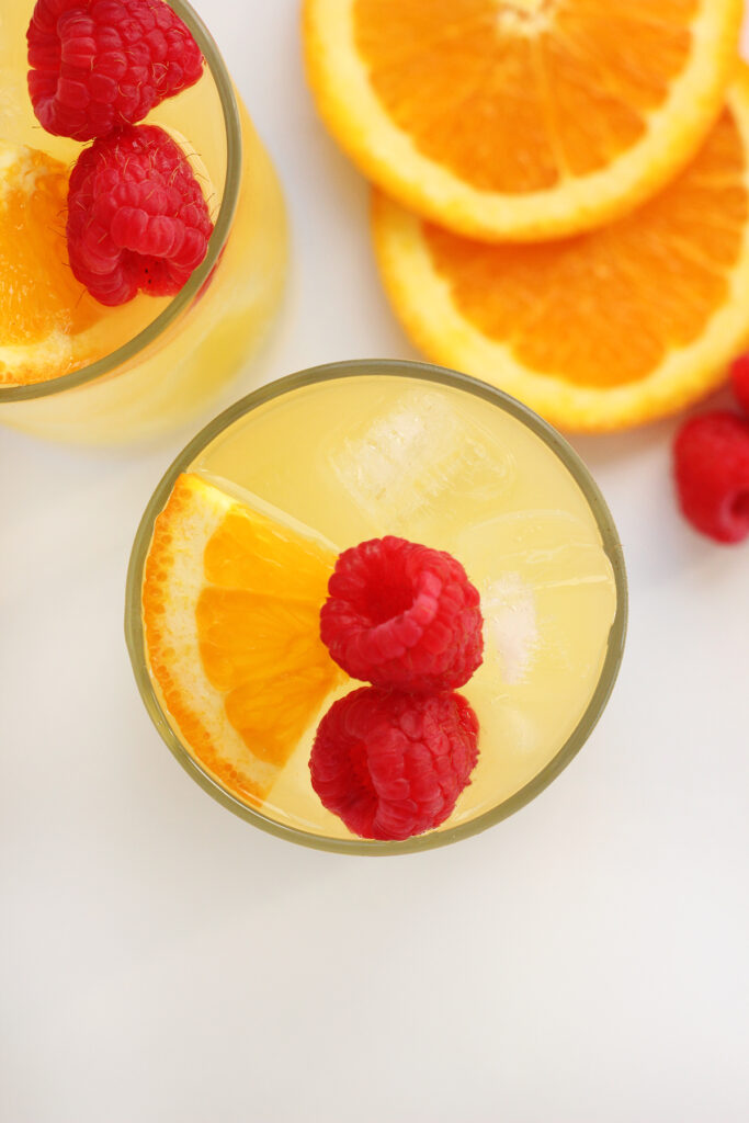 top down image focusing on the top of a glass filled with ice, two raspberries, and an orange slice. A second glass , orange slices, and raspberries are off to the side