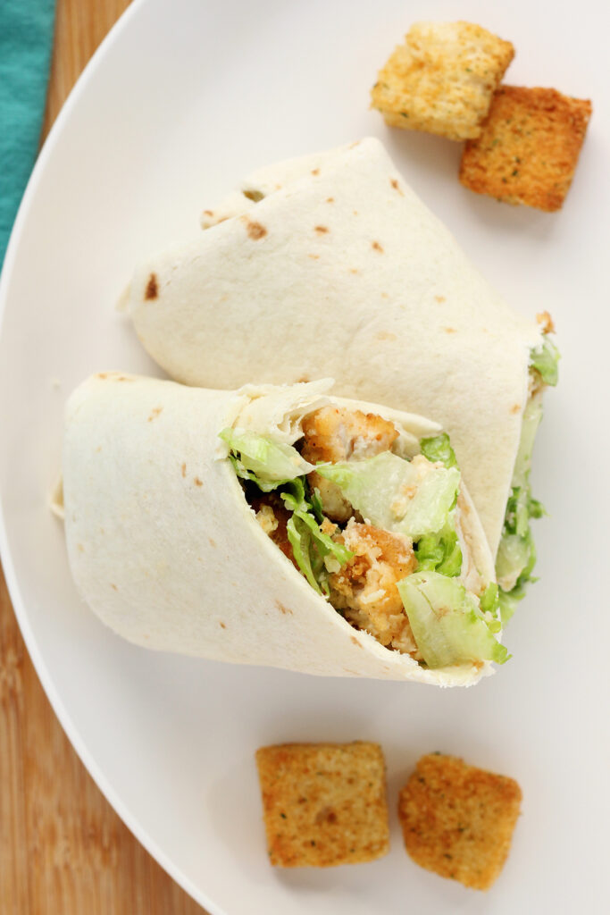 close up image showing a salad chicken wrap that has been sliced in half at a diagonal and is stacked on top of itself on a white plate with a few croutons around the wrap. 