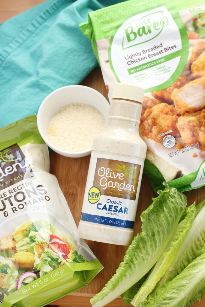 top down image showing a wooden cutting board loaded with ingredients for a chicken wrap. Ingredients include a bag of olive garden croutons, olive garden classic caesar dressing, pieces of romaine lettuce, a bowl of shredded parmesan cheese, and a bag of just bare chicken chunks with a teal napkin