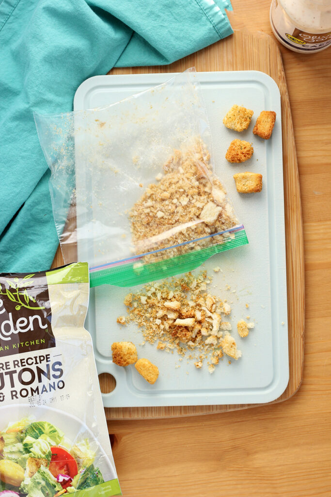 top down image showing a wooden cutting board on a wooden table. On top of that is another light blue cutting board with crushed croutons spilling out of a small ziplock bag. The original crouton bag is off to the side with a teal napkin