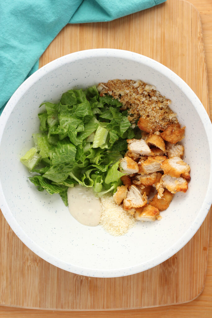top down image showing a white speckled mixing bowl filled with lettuce, chopped chicken, crushed croutons, parmesan cheese, and Caesar dressing. Ingredients are separated into their own portions.