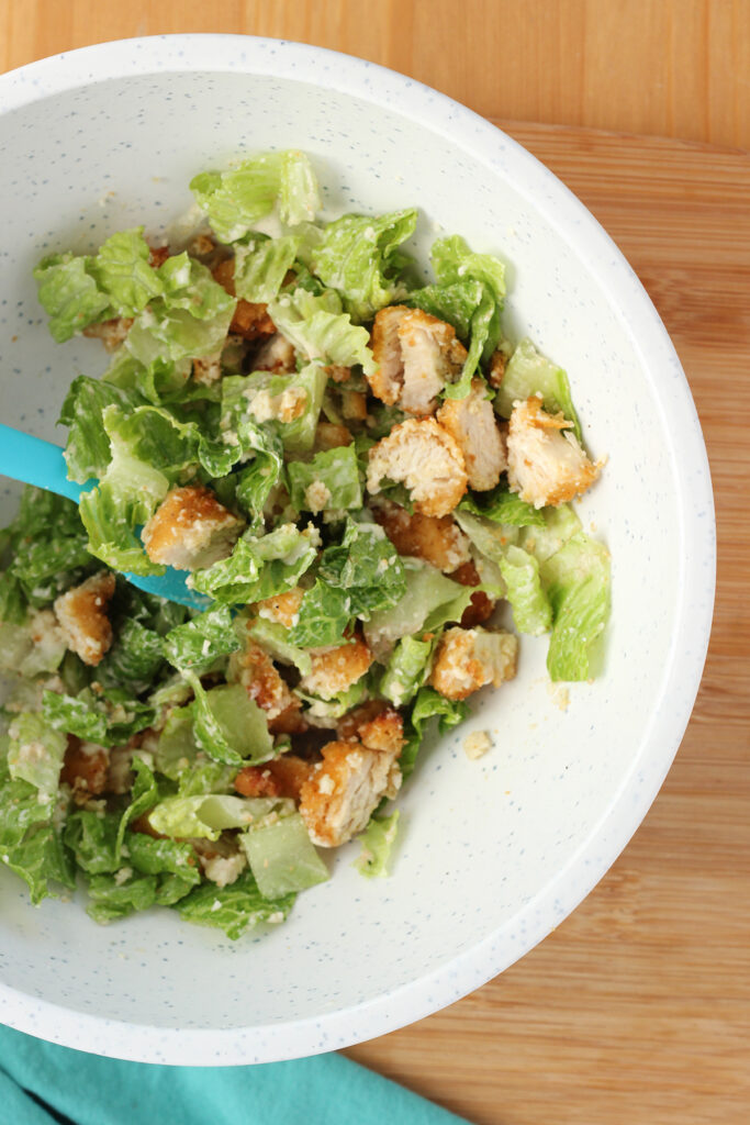 top down image showing a white speckled mixing bowl filled with lettuce, chopped chicken, crushed croutons, parmesan cheese, and Caesar dressing mixed together with a blue spatula