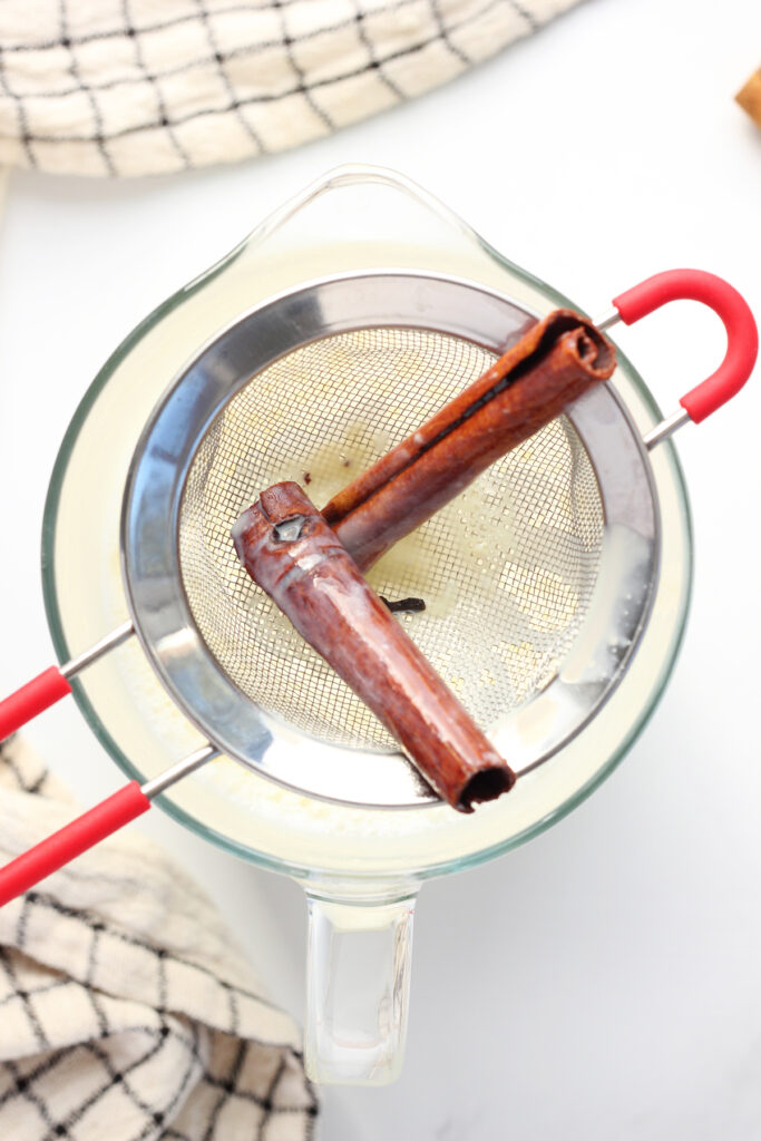 top down image showing a small strainer sitting on top of a pitcher.in the strainer are two cinnamon sticks and whole cloves
