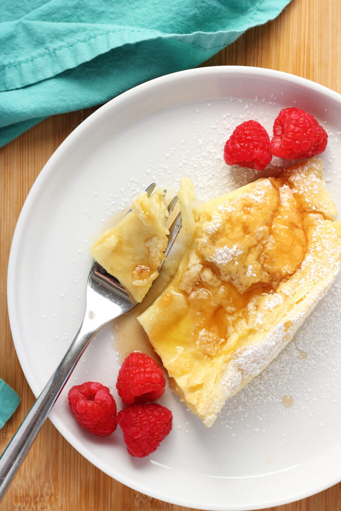 top down image showing a slice of dutch pancakes with a bite on a fork. The pancake has been dusted with powdered sugar with syrup drizzled on top. The pancake is sitting ona small white round plate with a teal napkin off to the side