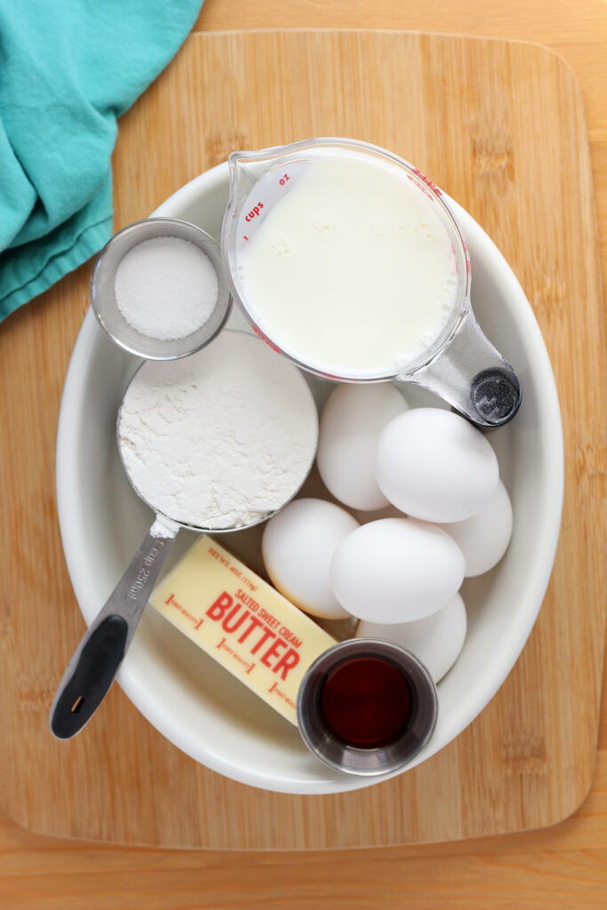 top down image showing a white oven baking dish filled with all of the ingredients needed to make a german pancake. It has six eggs, a stick of salted butter, vanilla extract, one cup of flour, one cup of milk, and sugar. The oval dish is sitting on a wooden table top with a teal napkin off to the side