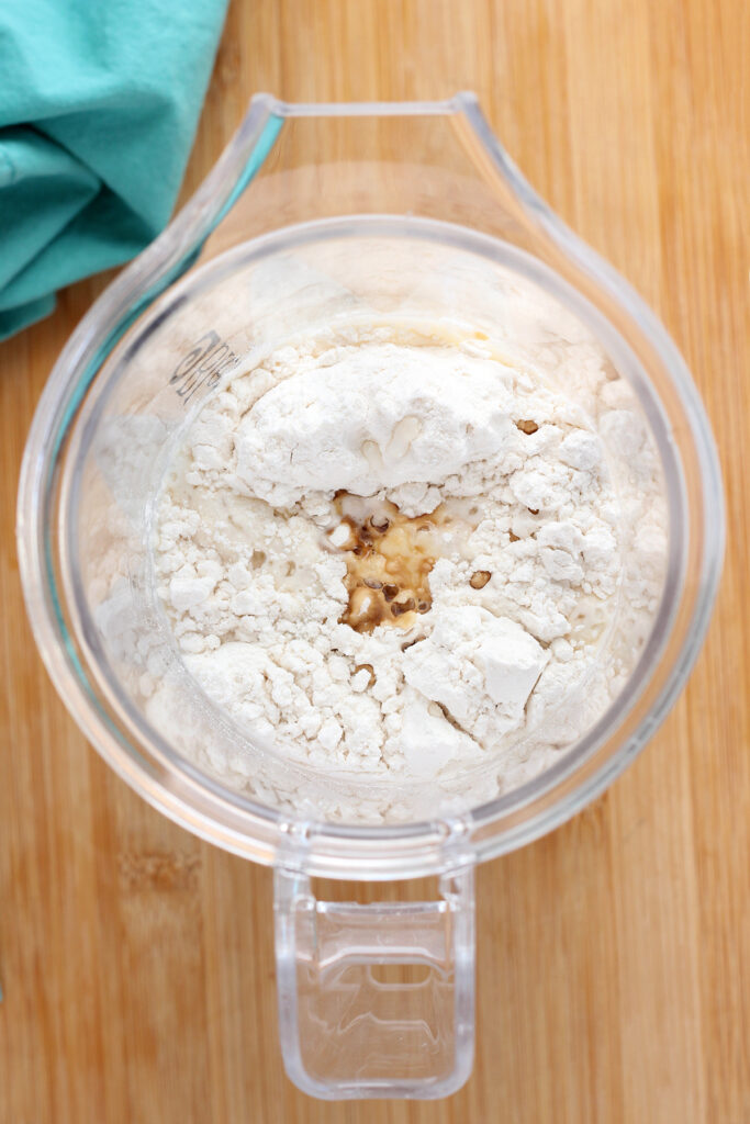 top down image showing a close up of a blender without the lid. Inside is milk, flour, and vanilla extract. The blender is sitting on a wooden table with a teal napkin