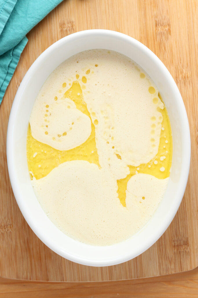 top down image showing a white baking dish that is sitting on a wooden table top with a teal napkin. Inside the dish is melted butter swirled with a pancake batter