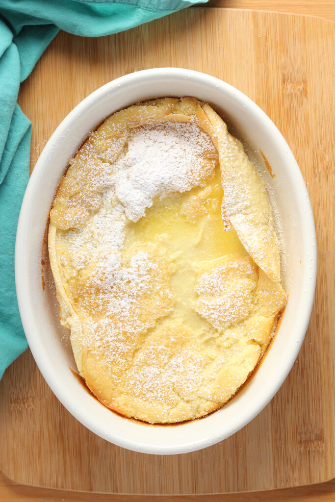 top down image showing a white baking dish that is sitting on a wooden table top with a teal napkin. Inside the dish is a baked pancake that has been dusted with powdered sugar