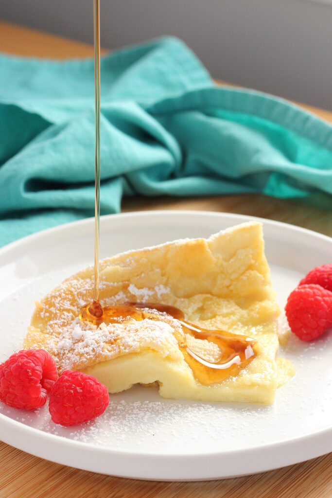 slice of a german pancake sitting on a small white round plate with raspberries dusted with powdered sugar. Syrup is being drizzled over the top