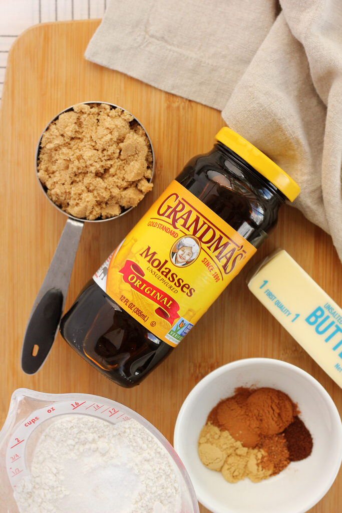 top down image showing a metal measuring cup filled with brown sugar, a jar of Grandma's Molasses, a stick of unsalted butter, a measuring cup filled with flour, and a small white bowl with various spices sitting on a wooden table top with a tan napkin