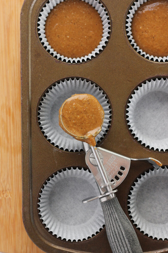 top down image showing a six cup cupcake tin sitting on a wooden table top. The cupcake tin is filled with paper liners that are being filled with a brown batter. Some batter is sitting inside a scoop that has been sitting on the tin