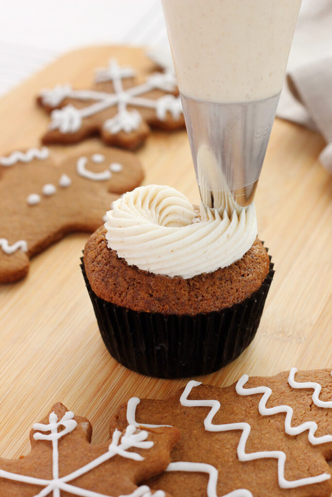 close up of a brown cupcake in a dark black wrapper being pipped with frosting. The cupcake is sitting on a wooden cutting board with gingerbread cookies off to the side. 