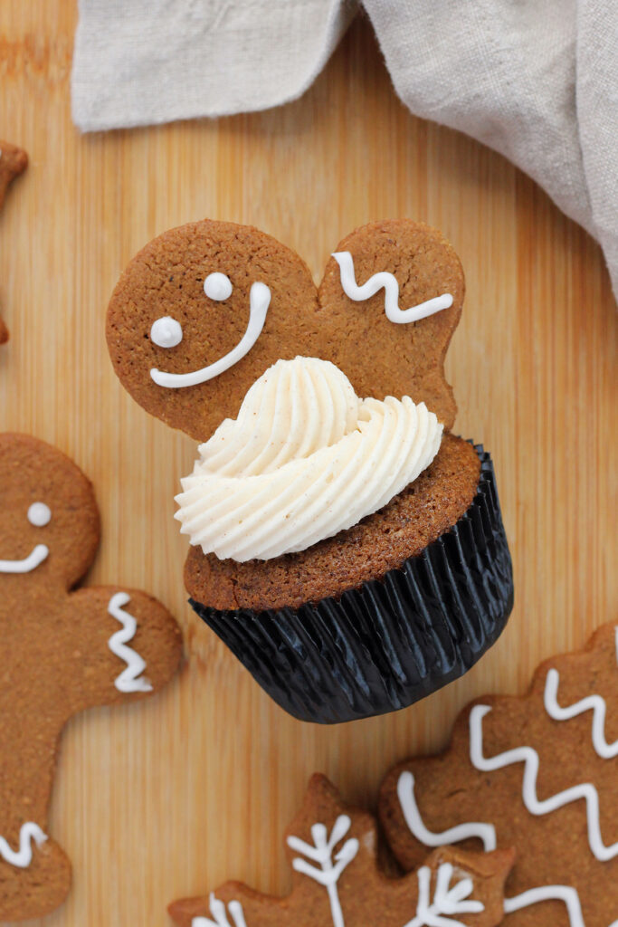 top down image showing a single cupcake in a black liner that is topped with a white frosting and half of a gingerbread man pushed into the top. More gingerbread cookies are around the sides with a tan napkin