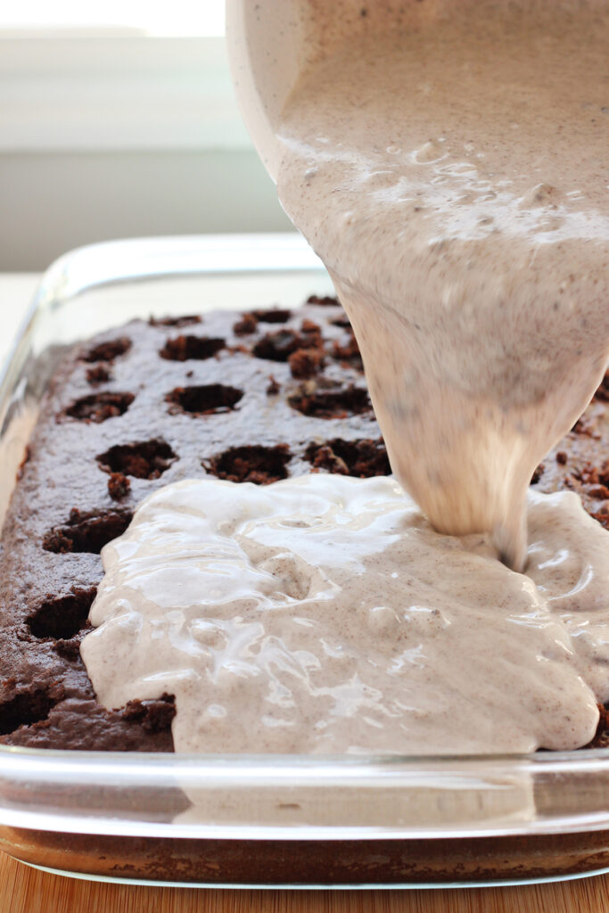 cookies and cream pudding being poured over the top of a baked chocolate cake in a glass pan that has been poked with holes all across the top